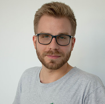 Mann mit Bart, Brille, lächelt leicht, trägt graues T-Shirt vor hellem Hintergrund.
