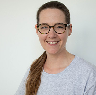 Person mit Brille lächelt freundlich, trägt ein graues Shirt, hat braune Haare und helle Haut.